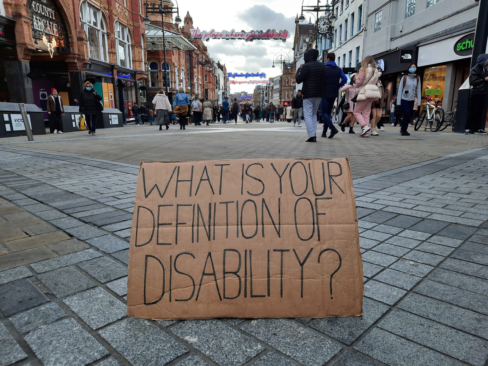 Image of a cardboard sign with the words on it ‘what is your definition of disability?’. The sign is on a pavement with shoppers walking in the background.