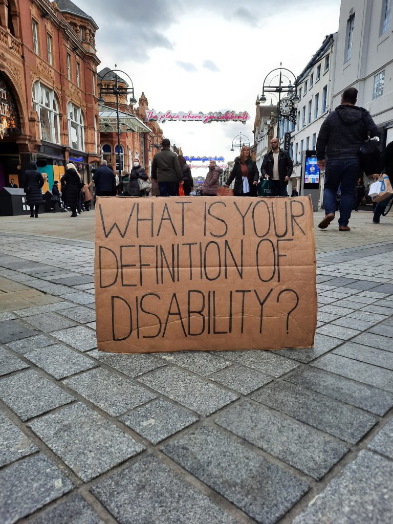 Image of a cardboard sign with the words on it ‘what is your definition of disability?’. The sign is on a pavement with shoppers walking in the background.