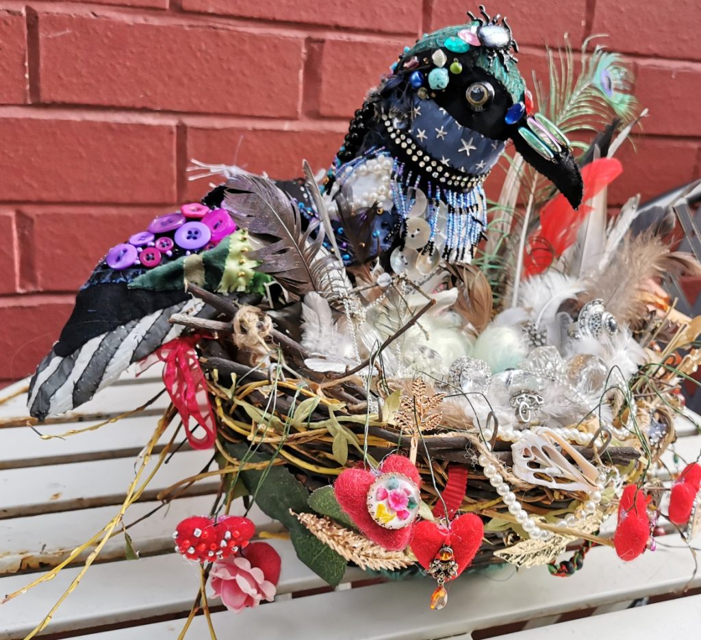 Image of a colourful bird sculpture, made from a variety of things, feathers, ribbons, broken jewellery and velvets, perched on a table, in front of a brick wall.