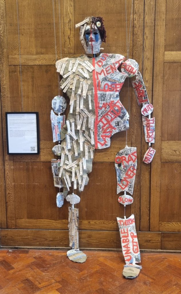 The artwork is a large hanging puppet structure. The right side is covered with hessian cloth and printed names of the females who were termed as ‘idiot’ or ‘imbecile’ in York Union Workhouse on census records for 1901. Down the middle of the puppet is a red painted line. On the left side, the puppet is covered with newspaper and red painted words such as ‘loser, loner, lazy’. On the head of the puppet, on the right side is a pin badge with a sunflower on it an the words ‘face mask exempt’.